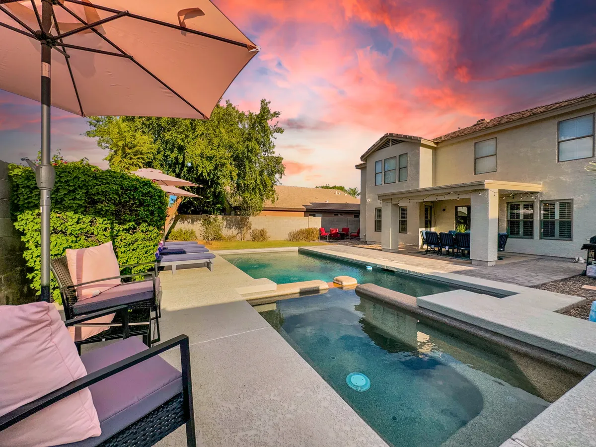 A tranquil exterior scene featuring a swimming pool, outdoor seating area with tables and pink umbrellas, and lush landscaped greenery. The setting is ideal for outdoor dining and social gatherings, especially in the evening or at sunset.