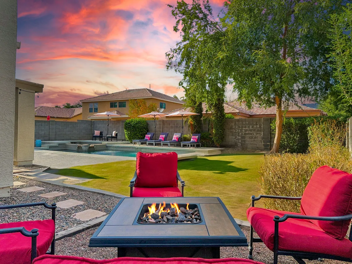 The scene features a tranquil backyard setting at sunset, with a fire pit surrounded by lounge chairs, the pool glistening in the background, and a cozy, welcoming mood perfect for evening gatherings. This area is staged for relaxation and conversation under the stars.