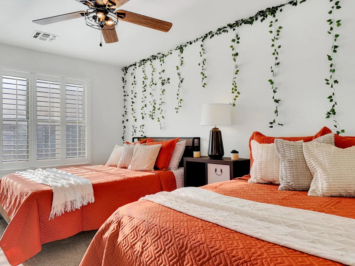 A cheerful bedroom with two beds featuring orange covers, green vine wall decor, a modern ceiling fan, and a shared nightstand. The space feels bright, inviting, and ideal for multiple guests or children.
