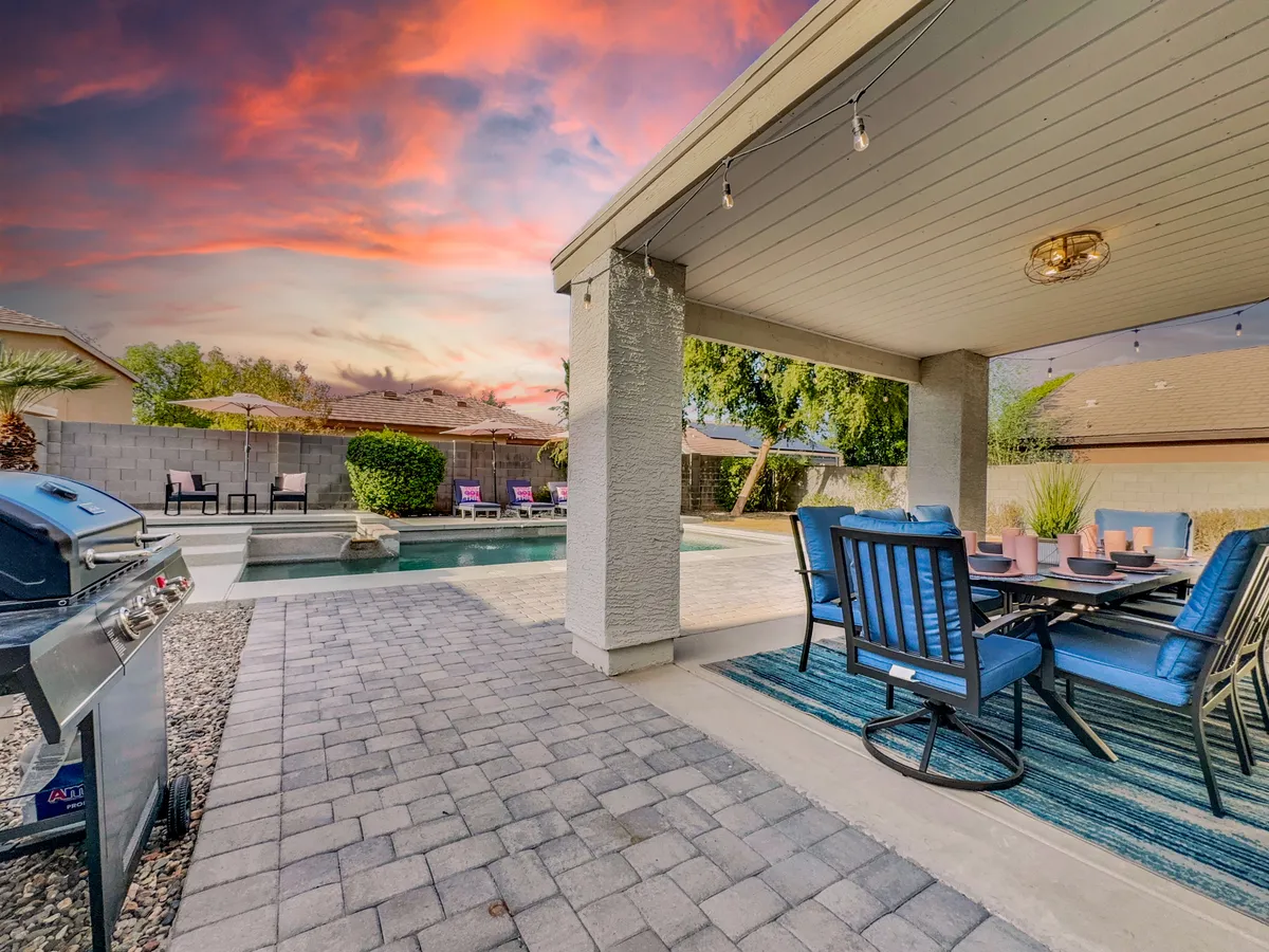 This image showcases the backyard pool area with a clear view of the inviting swimming pool, a dining area set up for outdoor meals, a barbecue grill, and an expansive sky above. The scene is arranged for casual poolside dining and relaxation, capturing a resort-style atmosphere.
