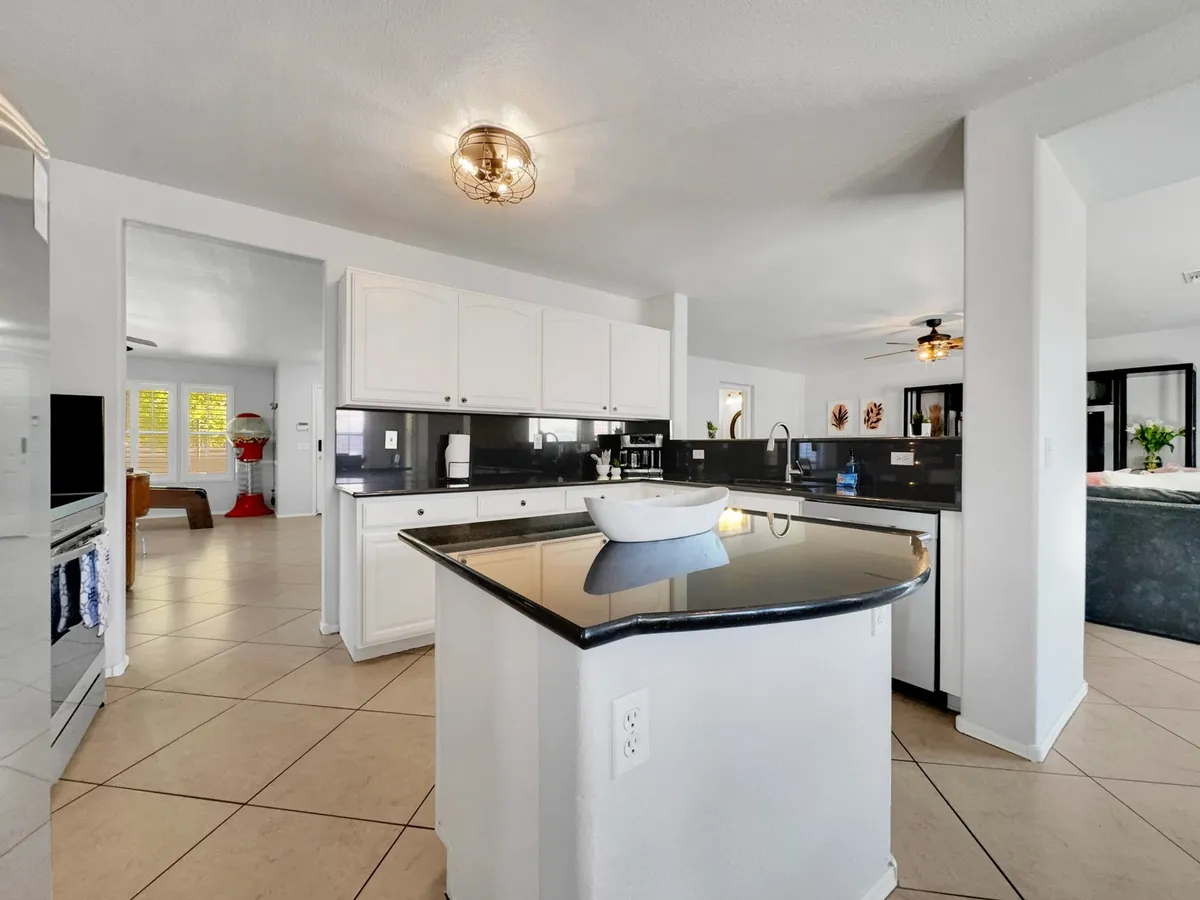 A bright, modern kitchen featuring a kitchen island, sleek black granite countertops, and white cabinetry. Sunlight pours in, enhancing the inviting atmosphere, and the space is clearly designed for both culinary activities and hosting. The layout supports seamless movement between cooking and socializing.