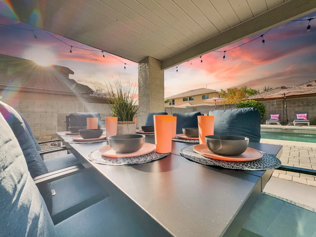 An inviting exterior dining table is set for a meal beside a sparkling swimming pool, with a beautiful sunset sky and festive string lights overhead. The atmosphere is tranquil yet lively, suggesting evenings out and social gatherings.