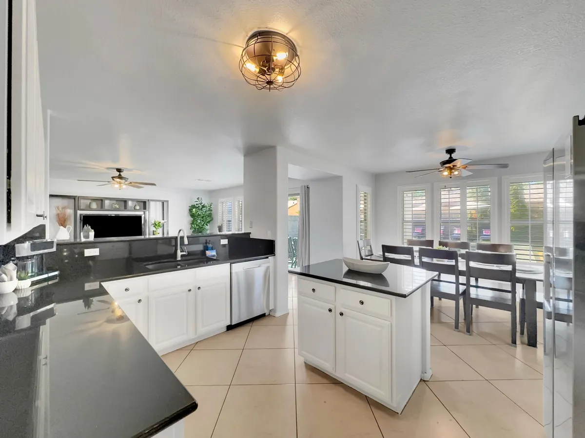 A view of the dining area with a large window, black granite countertops, white cabinets, and wooden chairs. The scene is bright and functional, ideal for both intimate family breakfasts and larger gatherings. The open flow between kitchen and dining is clearly depicted, highlighting the connection between cooking and sharing meals.