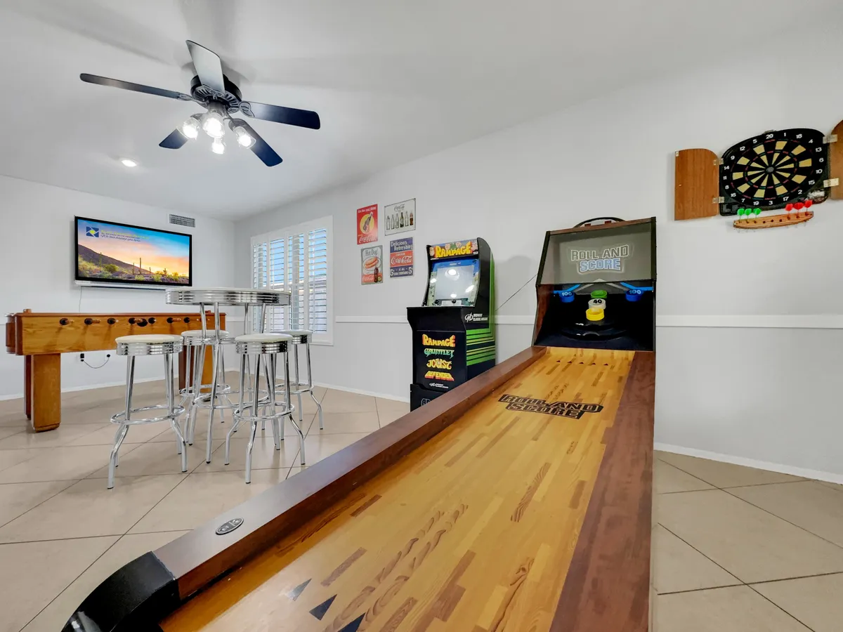 A vibrant, well-lit game room featuring a foosball table, arcade game machine, skee-ball machine, dartboard, and a TV with a scenic view in the background. The space appears active and playful, set up for group fun and lively game tournaments.