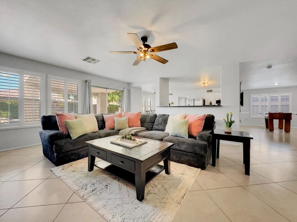 A modern living area featuring a large sectional sofa, coffee table, and decorative plants. Sunlight streams through expansive windows, bathing the room in natural light, while the layout encourages socializing and relaxation.